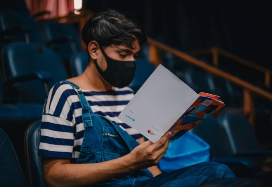 Foto de adolescente com máscara de proteção cobrindo a boca e o nariz sentado lendo uma cartilha, que ele segura com a mão direita. Na contracapa da publicação estão os logos do UNICEF e do Instituto BRF.